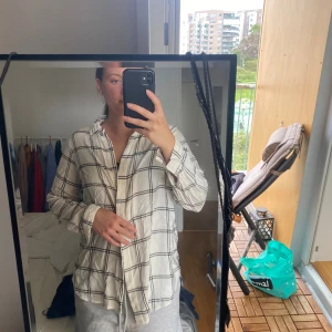 Vit rutig skjorta från H&M MAMA - Vit skjorta med svart rutigt mönster från H&M MAMA. Skjortan har klassisk krage, långa ärmar och knappar framtill. Tillverkad i mjuk bomull, perfekt för en chill och avslappnad stil.