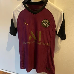 Vinröd PSG fotbollströja Jordan - Snygg PSG fotbollströja i samarbete med Jordan. Tröjan är vinröd med svarta och vita korta ärmar, guldiga detaljer och PSG-logga på bröstet. Materialet är lätt och andas. Liten i storleken