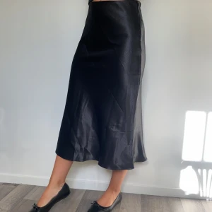 Svart satinlångkjol från SHEIN BAE - Supersnygg svart långkjol från SHEIN BAE i glansigt satinmaterial. Kjolen har en lätt utsvängd siluett och slät midja med dragkedja i sidan. Perfekt för dig som gillar stilrena och eleganta vibes. Passar dig som vill ha en chic look.