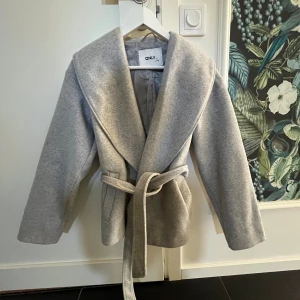 Ljusgrå omlottjacka från ONLY - Säljer en ljusgrå omlottjacka från ONLY med bred krage och bälte i midjan. Jackan har lång ärm och är tillverkad i ett mjukt material som känns både stilrent och bekvämt. Perfekt för lager-på-lager och snygg till jeans eller kjol.