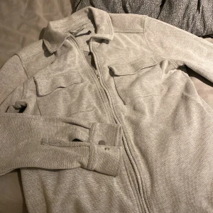 Grå overshirt från Dressmann, storlek S - Säljer en stilren grå overshirt från Dressmann i storlek S. Jackan har två bröstfickor med lock, klassisk krage och dragkedja framtill. Materialet är mjukt och känns som bomull, perfekt för lager-på-lager. Passar dig som gillar enkel och clean stil.