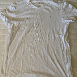 Vit t-shirt från GANT - Vit t-shirt från GANT med diskret logga på bröstet. Klassisk rund hals och korta ärmar. Tillverkad i mjuk bomull som känns skön mot huden. Perfekt basic-plagg med clean look och enkel passform.