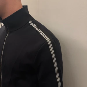 Svart Moncler kofta - Svart kofta från Moncler med vita ränder längs ärmarna och logotyp på axeln. Den har hög krage, hel dragkedja framtill. Perfekt för en sportig och clean stil.