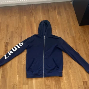 Marinblå hoodie från Zadig & Voltaire - Snygg marinblå hoodie från Zadig & Voltaire med vit text 'ZADIG' längs ena ärmen. Tröjan har dragkedja framtill, huva och två fickor. Fint skick och mycket lätt att stylea! Vid fler frågor/funderingar är det bara att skriva!