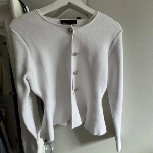 Vit kofta med glittriga knappar - Supersnygg vit kofta med långa ärmar och glittriga dekorativa knappar framtill. Modellen har en lätt figursydd passform och är tillverkad i ett stickat material som känns både mjukt och stilrent. Perfekt för dig som gillar en clean och chic look.