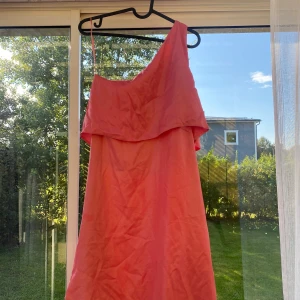 Korallrosa oneshoulder sommarklänning - Säljer en snygg korallrosa klänning med oneshoulder och volang upptill. Klänningen är kort och har en luftig passform, perfekt för varma dagar. Den har ett tunt axelband på ena sidan och är tillverkad i ett lätt material som känns skönt mot huden.
