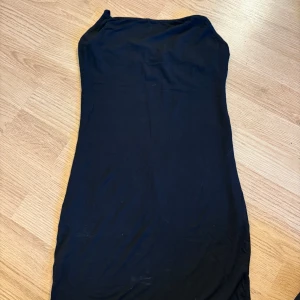 Svart lång klänning med smala axelband - En svart, tight långklänning med smala axelband som korsas i ryggen. Klänningen har en enkel och stilren design, perfekt för dig som gillar minimalistiskt mode. Materialet känns mjukt och stretchigt, vilket gör den bekväm att bära.