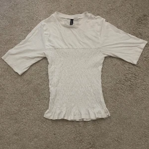 Vit smockad topp från Divided - Supersöt vit topp från Divided med smockad midja och volangkant nertill. Toppen har korta ärmar och rund halsringning. Perfekt för dig som gillar en feminin och trendig stil. Endast använd 4 gånger. Men har en liten fläck (sista bilden) men absolut inget man tänker på.