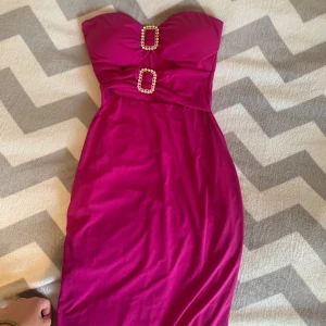 Lång cerise klänning med strassdetaljer - Säljer en snygg cerise långklänning med axelbandslös design och två glittriga strassspännen framtill. Klänningen har en figurnära passform och är perfekt för dig som vill sticka ut. Materialet känns mjukt och följsamt mot kroppen.