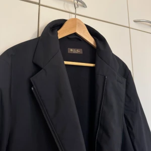 Navy rock från Loro Piana i kashmir - Säljer en navy rock från Loro Piana med klassisk krage och snygg brun mockadetalj på insidan av kragen. Rocken har dragkedja och knappar framtill, samt är tillverkad i en blandning av polyester och kashmir för en lyxig känsla. ✨ OBS: Jackan är navy (mörkblå), men kan se svart ut på bilderna.