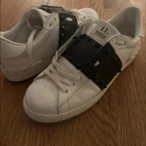 Valentino vita sneakers - Snygga vita Valentino Rockstud sneakers med svart band och ikoniska nitar över vristen. Skorna har rund tå, platt sula och klassisk snörning. Tillverkade i skinn med coola detaljer som ger en edgy vibe till din outfit. Storleken 43