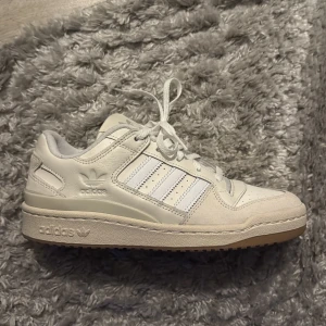 Adidas sneakers - Snygga vita Adidas sneakers med klassiska tre ränder på sidan och präglad logga. Skon har snörning, rund tå och en beige gummisula med mönster undertill. Ovandelen är i skinn och mocka för en clean och sportig look.