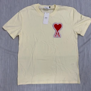 Gul t-shirt med hjärtdetalj från Ami - Snygg ljusgul t-shirt från Ami Alexandre Mattiussi med korta ärmar och rund hals. På bröstet sitter en stor röd och vit broderad patch med hjärta och bokstaven A. Tillverkad i mjuk bomull, perfekt för dig som gillar clean stil med en twist.