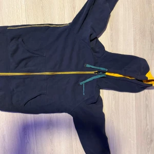 Mörkblå hoodie från Caterpillar - Snygg mörkblå hoodie från Caterpillar med gul dragkedja och gul detalj i huvan. Tröjan har långa ärmar, känguruficka och turkosa snören vid huvan. Perfekt för dig som gillar streetwear och vill sticka ut lite extra.