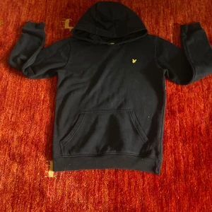 Svart hoodie från Lyle & Scott - Svart hoodie från Lyle & Scott med klassisk gul logga på bröstet. Tröjan har huva, känguruficka och långa ärmar. Perfekt för dig som gillar en enkel och stilren look. Mjuk och skön i bomullsmaterial.