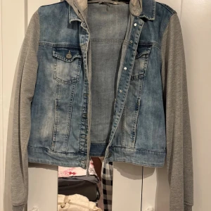 Jeansjacka med grå ärmar cropped - Säljer en snygg jeansjacka från LCW Jeans med ljusblå denim och grå sweatshirt-ärmar samt huva. Jackan har klassiska bröstfickor med knappar och en avslappnad streetstil. Perfekt för dig som gillar lager-på-lager och vill ha en chill look.