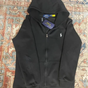 Svart hoodie från Polo Ralph Lauren - Svart hoodie från Polo Ralph Lauren med dragkedja och huva. Klassisk vit broderad logga på bröstet och ribbade muddar. Tröjan är i bomull och har en clean och stilren look som funkar till allt. Perfekt för dig som gillar enkel och snygg streetstil.