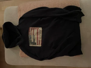 Mörkblå hoodie från Ralph Lauren - Svart hoodie från Ralph Lauren Denim & Supply med stor amerikansk flagga och logga på bröstet. Tröjan har huva och magficka, tillverkad i mjuk bomull. Perfekt för dig som gillar streetwear och snygga detaljer. Storleken är M men passar nån som vanligtvis har L