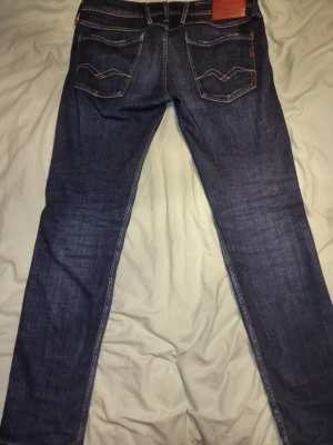 Replay jeans mörkblå tapered 33/32 - Snygga mörkblå jeans från Replay med klassisk femficksdesign och bruna läderdetaljer vid linningen. Tapered passform som sitter skönt och ger en modern siluett. Jeansen har kontrastsömmar och är tillverkade i slitstark denim.