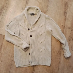 Stickad cardigan - Säljer en beige stickad kofta från H&M med grova kabelstickade detaljer och stora mörka knappar framtill. Koftan har en bred krage och långa ärmar med ribbade muddar. Perfekt för dig som gillar mysiga och stilrena plagg.skriv innan köp!