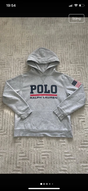 Grå hoodie Polo Ralph Lauren M - Snygg grå hoodie från Polo Ralph Lauren med stor logga på bröstet och amerikansk flagga på ena ärmen. Tröjan har huva, magficka och är fodrad med mjuk fleece. Perfekt för dig som gillar klassisk streetstyle och vill ha något bekvämt och stilrent.