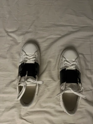 Valentino Garavani Open sneakers vit/svart - Säljer ett par snygga Valentino Garavani Open sneakers i vitt skinn med bred svart rem över mitten. Klassisk rund tå, platt sula och vita snören. Snygga nitar på hälen ger extra edge. Perfekt för dig som gillar lyxiga och stilrena sneakers.