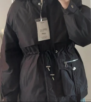 Vinterjacka / anorak - Snygg, funktionell och bekväm medellång vinterjacka för kvinnor med både justerbar midja och justerbar huva. Har både knappar och dragkedja & två stora fickor som det syns på bilden. Fuskpäls på insidan och det finns även borttagbar svart fuskpäls för huvan (meddela mig för bild). Ny med prislapp.För frågor eller fler bilder är det bara att höra av sig!