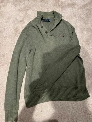 Olivgrön stickad tröja Polo Ralph Lauren - Snygg olivgrön stickad tröja från Polo Ralph Lauren med ribbad krage och två knappar vid halsen. Tröjan har långärmad passform och klassisk röd logobrodyr på bröstet. Perfekt för dig som gillar stilrena och tidlösa plagg.