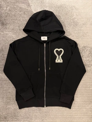 Svart hoodie med dragkedja från Ami - Svart hoodie från Ami Alexandre Mattiussi med stor vit och svart hjärtpatch på bröstet. Tröjan har huva med snörning, två fickor fram och hel dragkedja. Materialet är mjuk bomull och passformen är normal. Perfekt för dig som gillar stilrena streetwear-plagg.