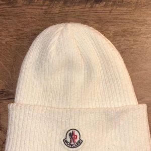 Vit ribbad Moncler mössa - Snygg vit ribbstickad mössa från Moncler med klassisk logotyp framtill. Mössan har uppvikt kant och är tillverkad i mjukt stickat material som håller dig varm och stilren under kalla dagar. Perfekt för dig som vill ha en clean look med exklusiv känsla.