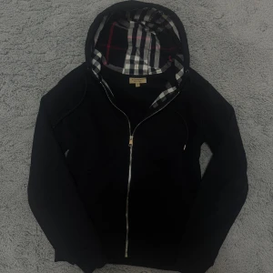 Svart Burberry zip hoodie - Svart hoodie från Burberry i storlek M med klassiskt rutigt foder i huvan och snyggt broderad logga på bröstet. Tröjan har hel dragkedja framtill, snörning i huvan och långa ärmar. Perfekt för dig som gillar stilrena och exklusiva plagg.