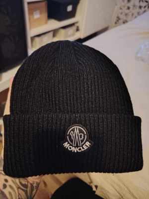 Svart ribbstickad mössa från Moncler - Svart ribbstickad mössa från Moncler med uppvikt kant och broderad logotyp framtill. Klassisk och stilren design som passar perfekt till kyliga dagar. Materialet känns mjukt och värmande.