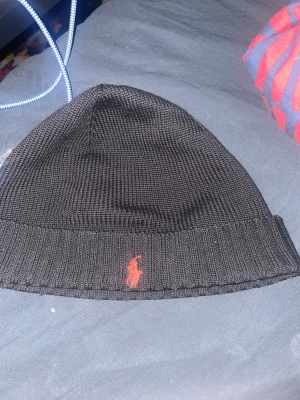 Mössa från Polo Ralph Lauren - Svart stickad mössa från Polo Ralph Lauren med uppvikt kant och röd broderad logotyp framtill. Tillverkad i 100% ull för extra värme och komfort. Klassisk och stilren design som passar till många olika outfits. Lite använd men märks inget bara på insidan