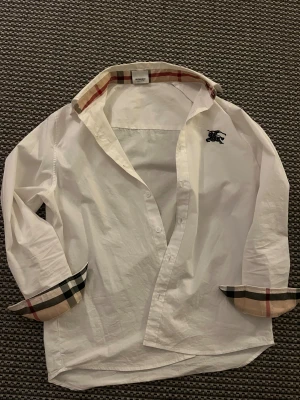 Vit Burberry skjorta med rutig detalj - Snygg vit skjorta från Burberry med klassiska rutiga detaljer på krage och manschetter. Skjortan har lång ärm, knappar framtill och Burberrys broderade logga på bröstet. Tillverkad i mjuk bomull och har en loose passform som ger en chill vibe.