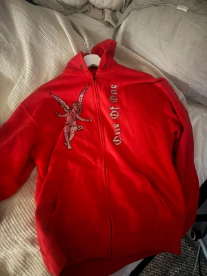 Röd One of One Hoodie - Säljer en röd hoodie med dragkedja från Love Of One. På framsidan finns en stor ängel i strass och texten 'Love Of One' i glittriga bokstäver. Tröjan har huva och fickor framtill, perfekt för en cool streetstil.