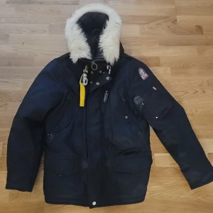 Svart parkajacka S - Svart parkajacka vit päls på huvan och gul detalj vid halsen. Jackan har flera fickor, dragkedja och knappar framtill, samt broderad logga på ärmen. Insidan är quiltad med orange detaljer. Slim fit-modell, perfekt för kalla vinterdagar. Akopia