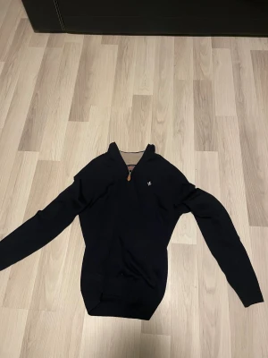 Svart half zip stickad tröja morris - Svart stickad tröja från Gant med half zip och liten broderad logga på bröstet. Tröjan har lång ärm och ribbade muddar vid ärmslut och nederkant. Perfekt för dig som gillar klassisk stil med en modern touch. Tar emot byten 