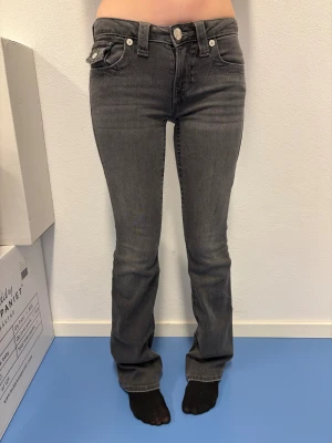 Grå bootcut jeans från True Religion - Snygga grå jeans från True Religion med bootcut passform och klassiska fickor med silverfärgade knappar. Jeansen har låg midja och detaljerade sömmar på bakfickorna. Perfekt för dig som gillar en cool och tidlös look.