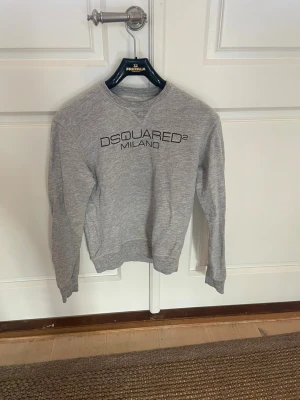Grå sweatshirt från Dsquared2 Milano - Tjena! Säljer en snygg grå sweatshirt från Dsquared2 Milano med svart logotyptryck på bröstet. Tröjan har rund hals, långa ärmar och ribbade muddar. Perfekt för dig som gillar stilrena och trendiga plagg. Skick: 8/10. Storlek: xs