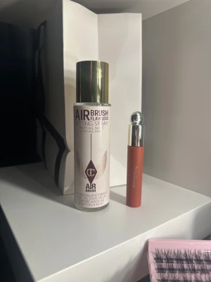 Charlotte Tilbury setting spray & Rare Beauty läppstift - Säljer en Airbrush Flawless Setting Spray från Charlotte Tilbury med guldfärgat lock och en matt röd läppstift från Rare Beauty med silvrigt lock. Perfekt för att fixera sminket och ge snygga läppar. Båda produkterna har stilren design.