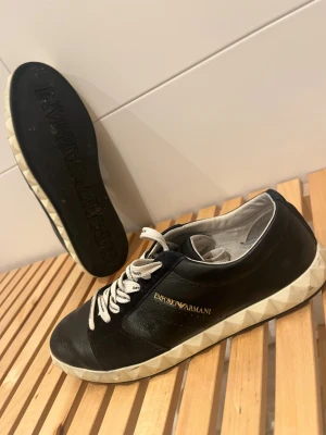 Svarta sneakers från Emporio Armani - Snygga svarta sneakers från Emporio Armani med vit, mönstrad sula och vita snören med logotyp. Skorna är tillverkade i skinn och har en rund tå. Guldtext med varumärket på sidan ger en lyxig touch. Perfekta för dig som gillar stilrena och exklusiva sneakers. Använda ett fåtal gånger