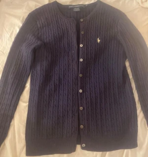 Marinblå kofta från Ralph Lauren Sport - Snygg marinblå kofta från Ralph Lauren Sport i 100% bomull. Kabelstickad design med långa ärmar, rund hals och knappar hela vägen. Denr i storlek M men passar även S. Pris kan diskuteras vid snabba köp. Den är nästan inte använd och inga slitage syns eller finns på koftan.