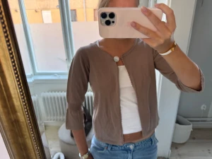 Beige kofta med knapp och volangkant - Säljer en beige kofta med trekvartsärm och söt volangkant vid ärmslut och halsringning. Koftan har en enkel knapp framtill och är gjord i mjukt stickat material. Perfekt att slänga över en topp för en chill look.