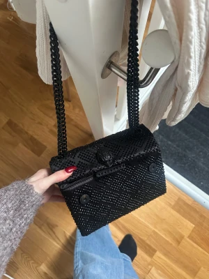 Svart axelväska Kurt Geiger med kedja - Snygg svart axelväska från Kurt Geiger, modell The Kensington. Väskan har glittriga svarta stenar i ett rutigt mönster och en grov kedja som axelrem. Materialet är satin och väskan stängs med magnetknappar. Perfekt för att lyfta din outfit.