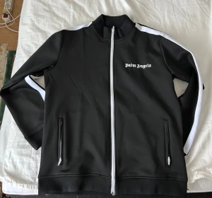Svart Palm Angels track jacket L - Palm Angels svart track jacket med vita ränder längs ärmarna och vit dragkedja. Jackan har ribbad krage, två fickor med dragkedja och broderad logga på bröstet. Materialet är 100% polyester och modellen är normal passform.