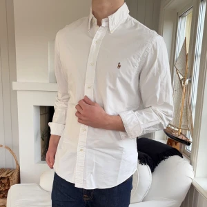 Vit slim fit skjorta från Ralph Lauren slim fit - Vit skjorta från Ralph Lauren i slim fit-modell med klassisk krage och knappar framtill. Skjortan har det ikoniska broderade Polo-logot på bröstet och är tillverkad i mjuk bomull. Långärmad och stilren design som passar perfekt till jeans eller chinos.