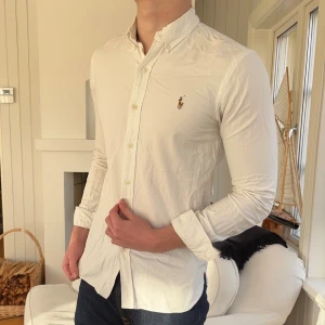Vit skjorta från Polo Ralph Lauren slim fit - Vit skjorta från Polo Ralph Lauren i slim fit med klassisk krage och knappar hela vägen. Liten broderad logga på bröstet. Skjortan är långärmad och har en stilren, tidlös look som passar till jeans eller chinos. Materialet är mjuk bomull som känns skönt mot huden.