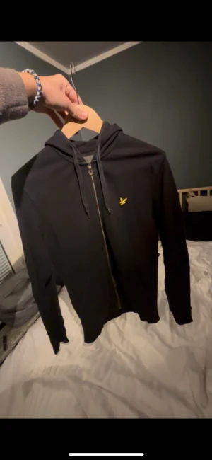 Svart hoodie från Lyle & Scott - Svart hoodie från Lyle & Scott med gul broderad logga på bröstet. Tröjan har huva med snörning, hel dragkedja framtill och långa ärmar. Materialet är mjuk bomull och passformen är normal. Perfekt för chill dagar och enkel att matcha med jeans eller joggers. Storlek xs