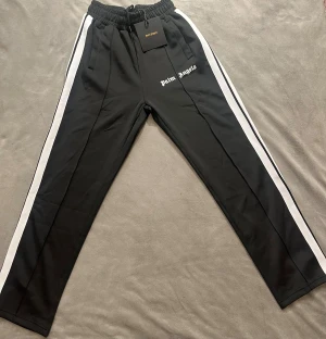 Palm Angels svarta mjukisbyxor med vita ränder - Svarta mjukisbyxor från Palm Angels med vita ränder längs sidorna och logotypen tryckt på vänster ben. Byxorna har elastisk midja med snörning och raka ben. Materialet är mjukt och har en lätt glansig finish, perfekt för en chill och sportig look.