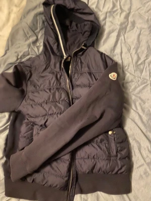 Mörkblå  Moncler cardigan  - Snygg mörkblå cardigan från Moncler med huva och dragkedja. Jackan har quiltad framsida, ribbade muddar och två fickor med tryckknappar. Moncler-logga på ärmen ger extra stilpoäng. Perfekt för dig som gillar streetwear och exklusiva märken. Storlek är 164 men passar mig som är 170 den är äkta och är köpt från sequro fashion äkta heta intyg kan lösas vid frågan